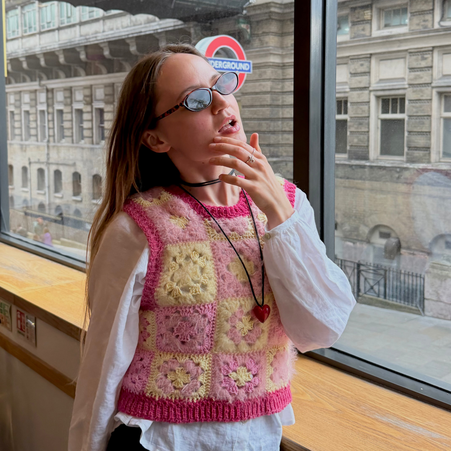 Woman wearing a colorful knitted vest and sunglasses, standing in front of a window with a cityscape view.