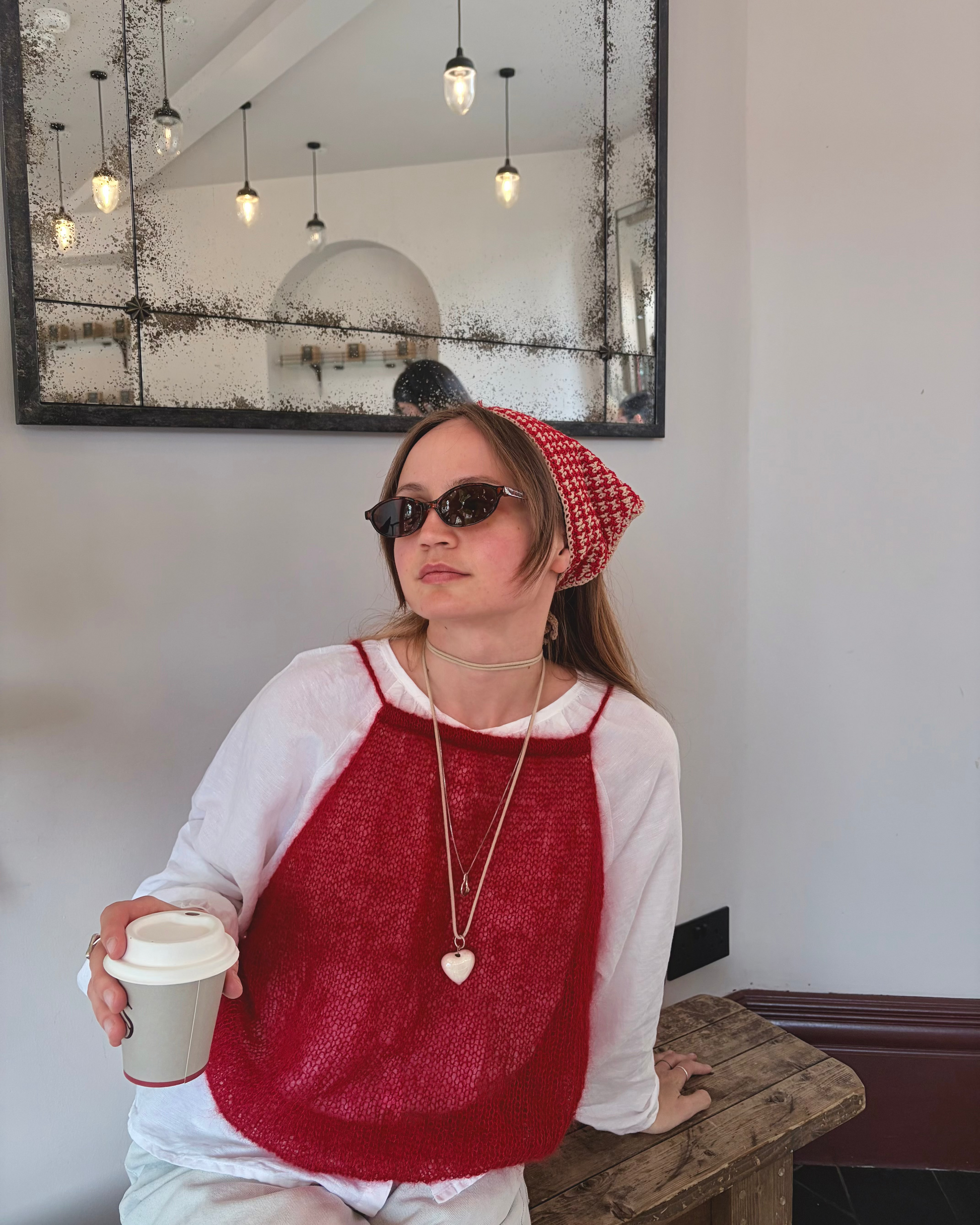 Woman wearing a red sweater and sunglasses, holding a coffee cup indoors.