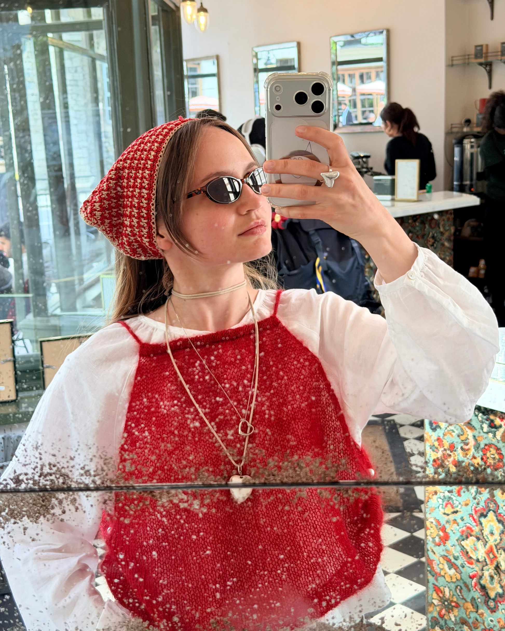 Woman taking a mirror selfie wearing a red knitted beanie and sunglasses indoors.